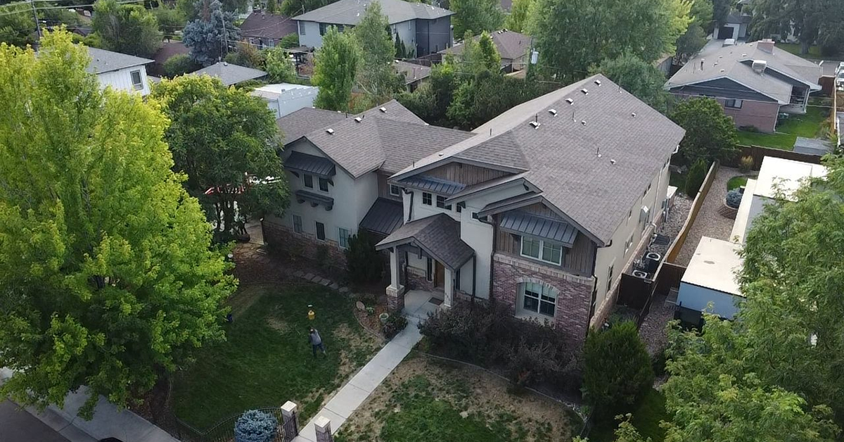 Aerial view of a two-story house with a gray roof, light-colored walls, and a brick facade. A person is visible in the yard.